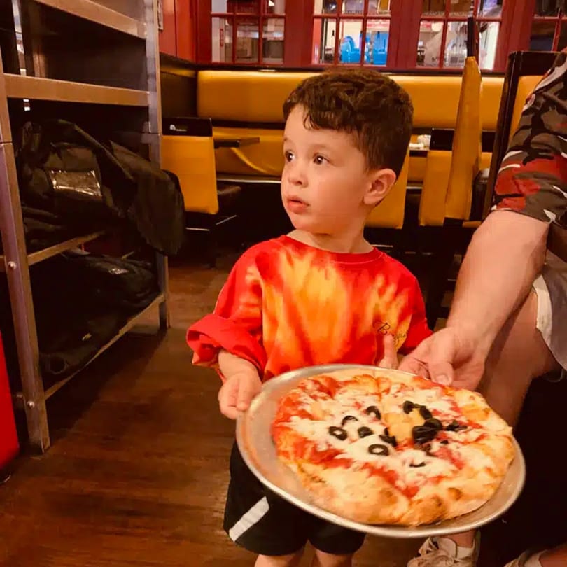 A young boy in a tie-dye shirt holds a small pizza with black olives at Biaggio Pizzeria.
