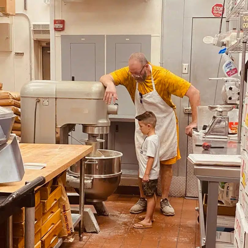 A man and a boy in a commercial kitchen, watching a large mixer, possibly making pizza dough.