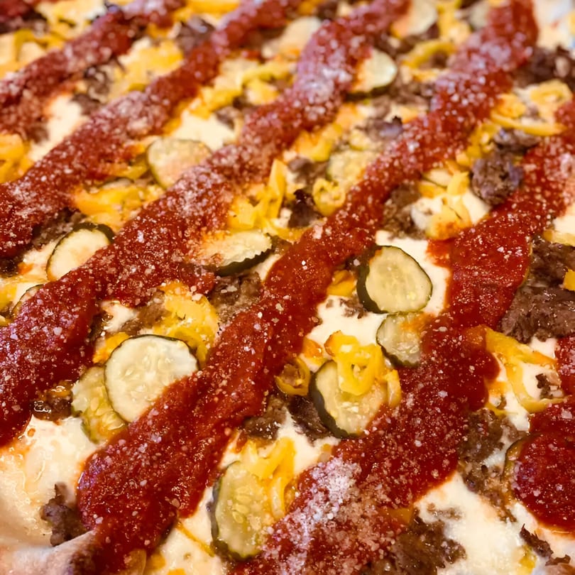 Close-up of a Biaggio Pizzeria pizza with ground meat, pickles, banana peppers, and ketchup drizzle.