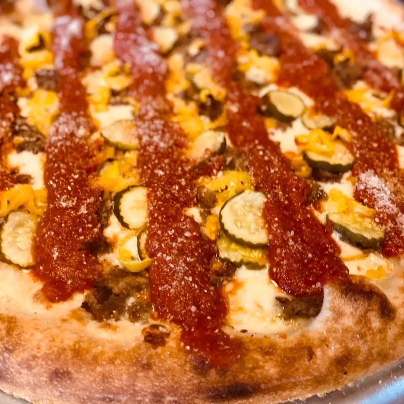 Close-up of a Biaggio Pizzeria pizza with pickles, ground meat, cheese, and red sauce drizzle.